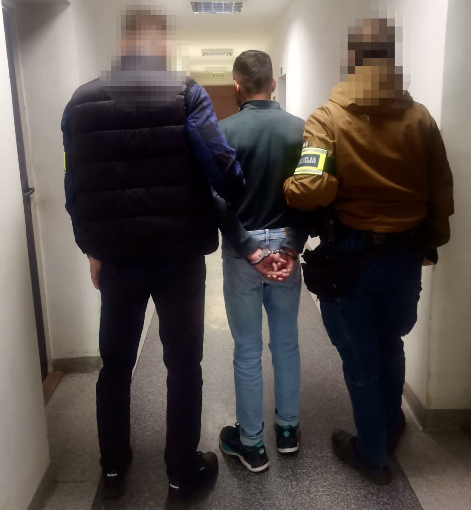 Mężczyzna z rękami skuty w kajdankach prowadzony przez dwóch funkcjonariuszy w korytarzu.