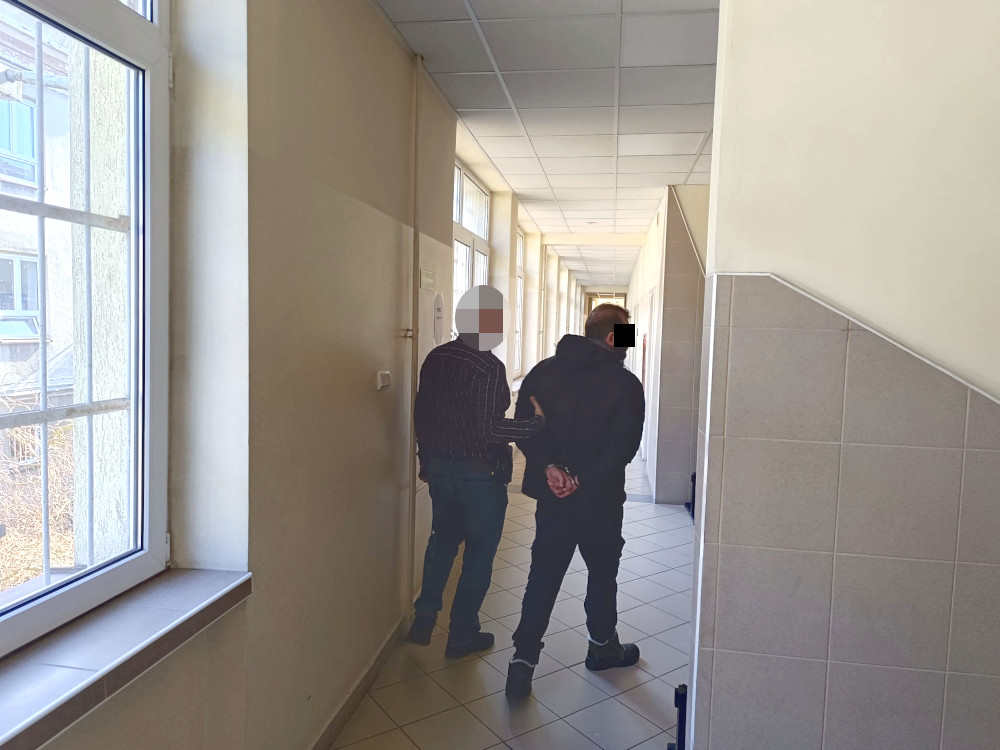 Mężczyzna w koszuli w kratę jest prowadzony przez funkcjonariusza w korytarzu budynku.