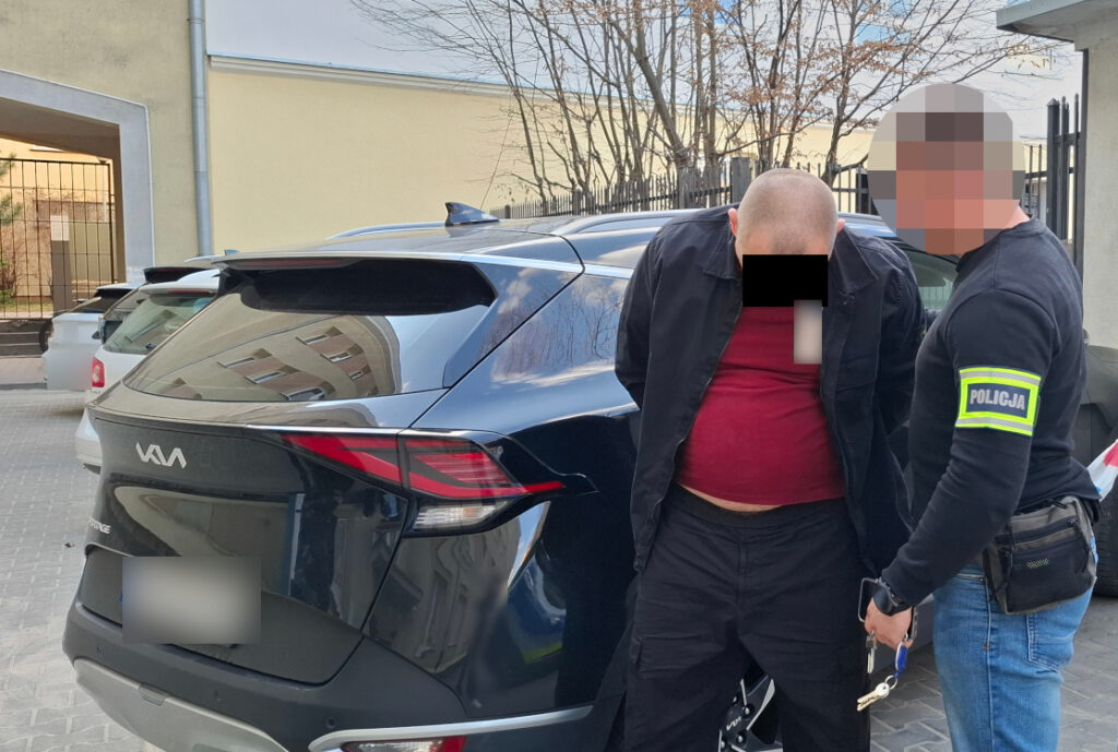 Mężczyzna w czerwonej koszulce jest zatrzymywany przez policjanta na parkingu, obok czarnego SUV-a.