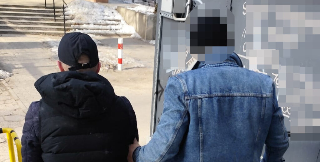 Mężczyzna w czapce i kurtce z kapturem stoi z tyłu, trzymając się za rękę z mężczyzną w niebieskiej kurtce dżinsowej, na tle schodów.