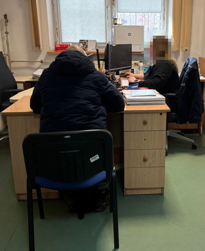 Mężczyzna w ciemnej kurtce siedzi przy biurku, rozmawiając z kobietą w czarnym ubraniu, pracującą przy komputerze.