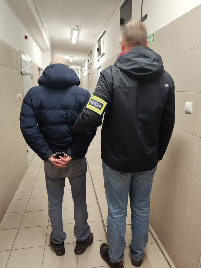 Mężczyzna w kajdankach prowadzony przez policjanta w korytarzu budynku.