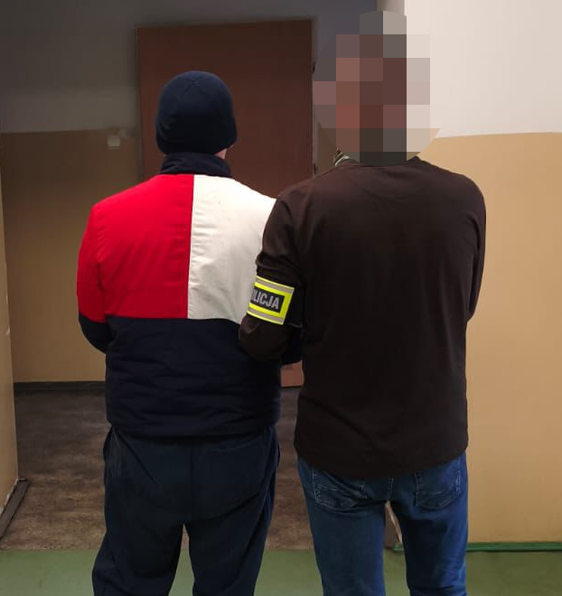 Mężczyzna w niebieskiej czapce i kolorowej kurtce stoi obok mężczyzny w brązowej bluzie z opaską "POLICJA" na ramieniu.