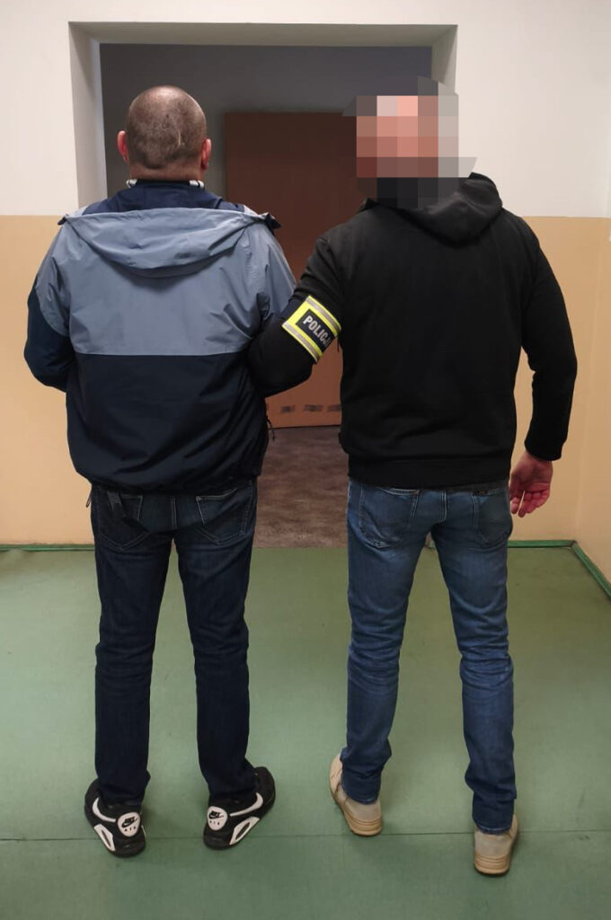 Mężczyzna w dżinsach prowadzony przez policjanta z opaską „POLICJA” w korytarzu budynku.