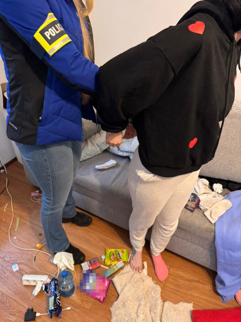 Policjantka zakłada kajdanki na ręce kobiety w czarnej bluzie z czerwonymi sercami w nieuporządkowanym pomieszczeniu.