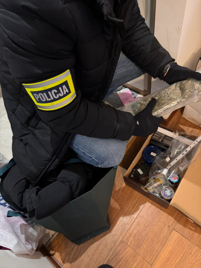 Funkcjonariusz policji w rękawiczkach trzyma zapakowaną substancję w pomieszczeniu z różnymi przedmiotami.