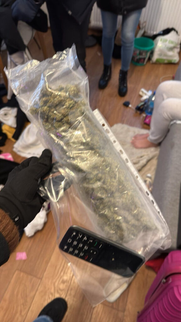 Mężczyzna w rękawiczkach trzyma przezroczystą torbę z marihuaną oraz pilot do telewizora, w tle nieuporządkowane pomieszczenie.