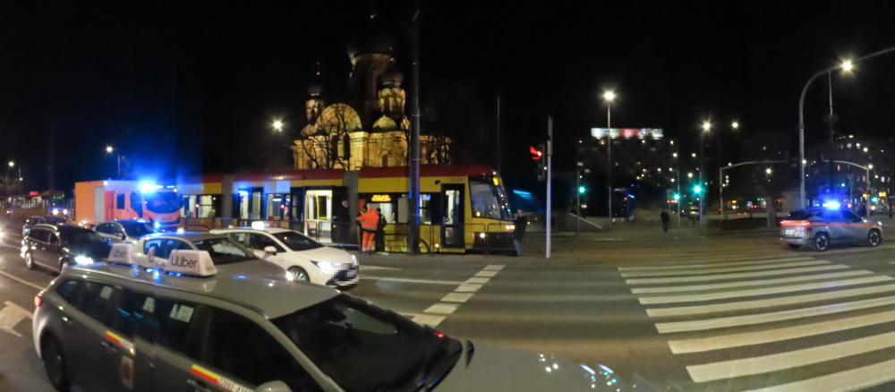 Nocna scena na ulicy z tramwajem zatrzymanym na przystanku, w tle oświetlona katedra i radiowóz z włączonymi sygnałami.