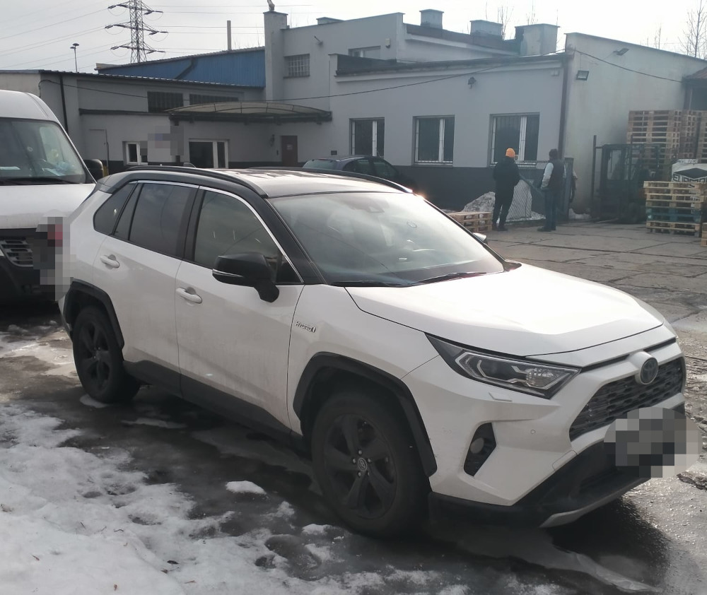 Biały SUV zaparkowany na terenie przemysłowym, w tle widoczni pracownicy oraz palety z towarem.