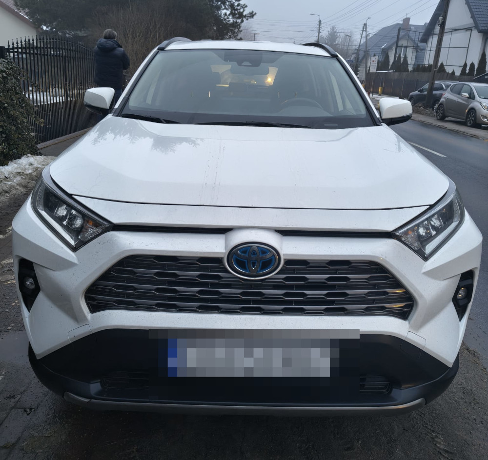 Biały SUV zaparkowany na ulicy, z widocznymi detalami przedniego grilla oraz osobą stojącą w tle.