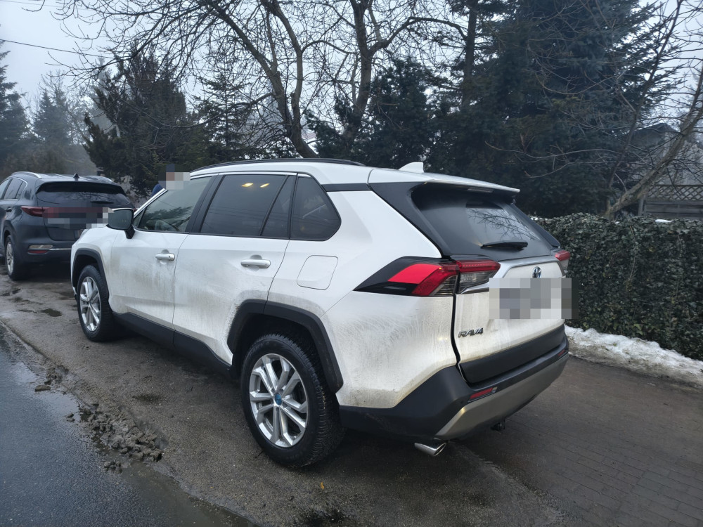 Biały SUV zaparkowany na wąskiej ulicy w otoczeniu drzew i krzewów, z widocznym brudem na karoserii.