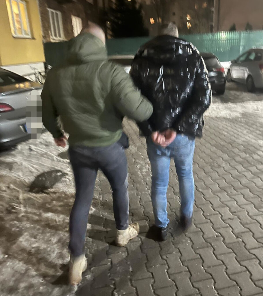 Mężczyzna w zielonej kurtce prowadzi drugiego mężczyznę w czarnej kurtce, idąc po wyłożonym kostką chodniku.