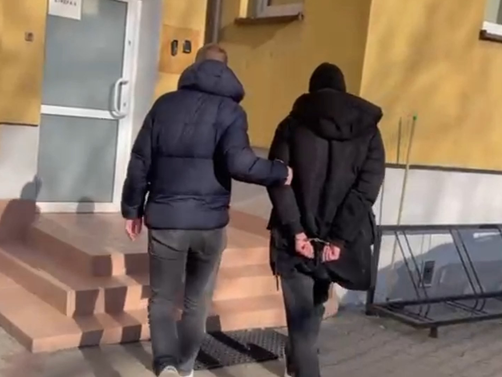 Mężczyzna w ciemnej kurtce prowadzi osobę w kajdankach w stronę drzwi budynku.