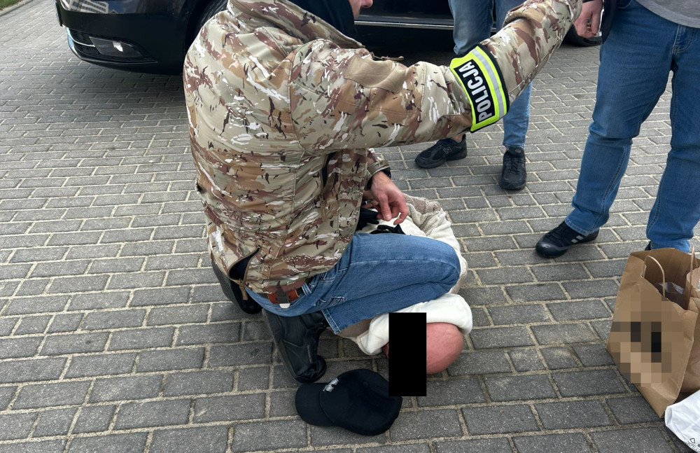 Policjant w kamizelce taktycznej przeszukuje osobę siedzącą na chodniku obok zaparkowanego samochodu.