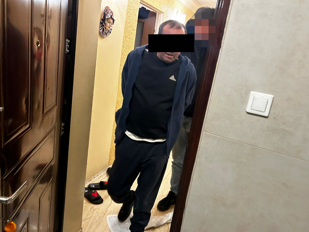 Mężczyzna w dresie wychodzi z mieszkania, podczas gdy inna osoba stoi w tle, w wąskim korytarzu.