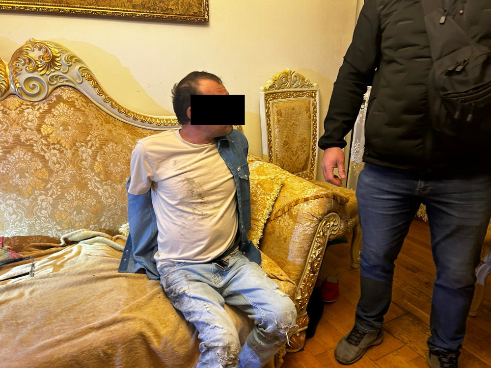 Mężczyzna w rozciągniętej koszulce siedzi na kanapie w eleganckim wnętrzu, obok stoi inny mężczyzna w czarnej kurtce.