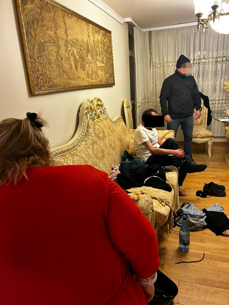 Mężczyzna w czarnej kurtce stoi obok siedzącego mężczyzny w białej koszulce, w eleganckim salonie z ozdobnym obrazem na ścianie.