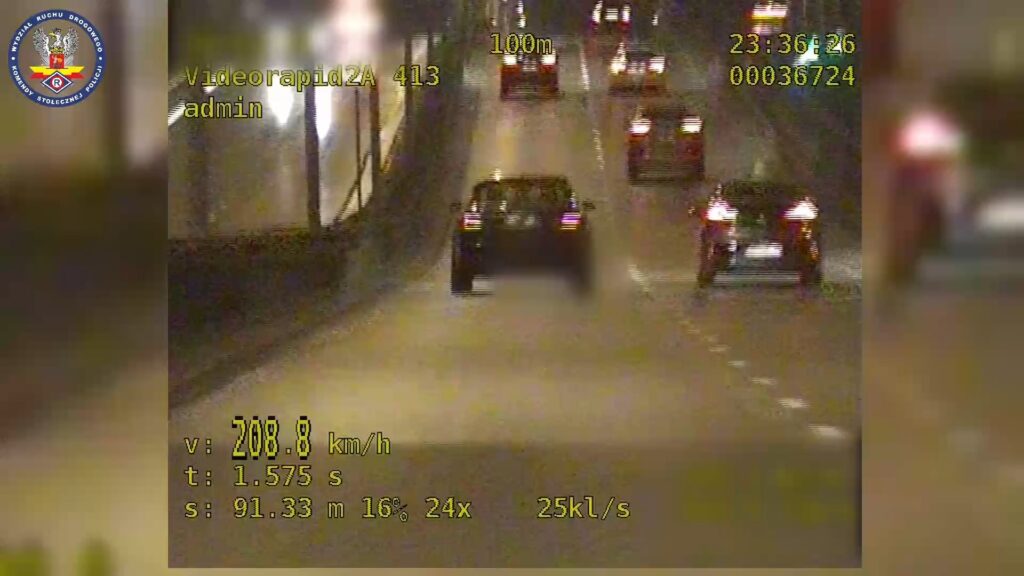 Samochód poruszający się z prędkością 208,8 km/h na oświetlonej drodze, z innymi pojazdami w tle.