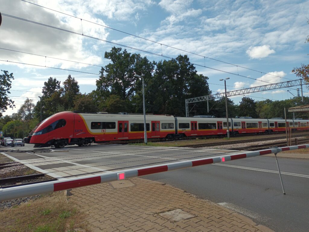 Pociąg SKM przejeżdża przez przejazd kolejowy, a w tle widoczne są drzewa i niebo z chmurami.