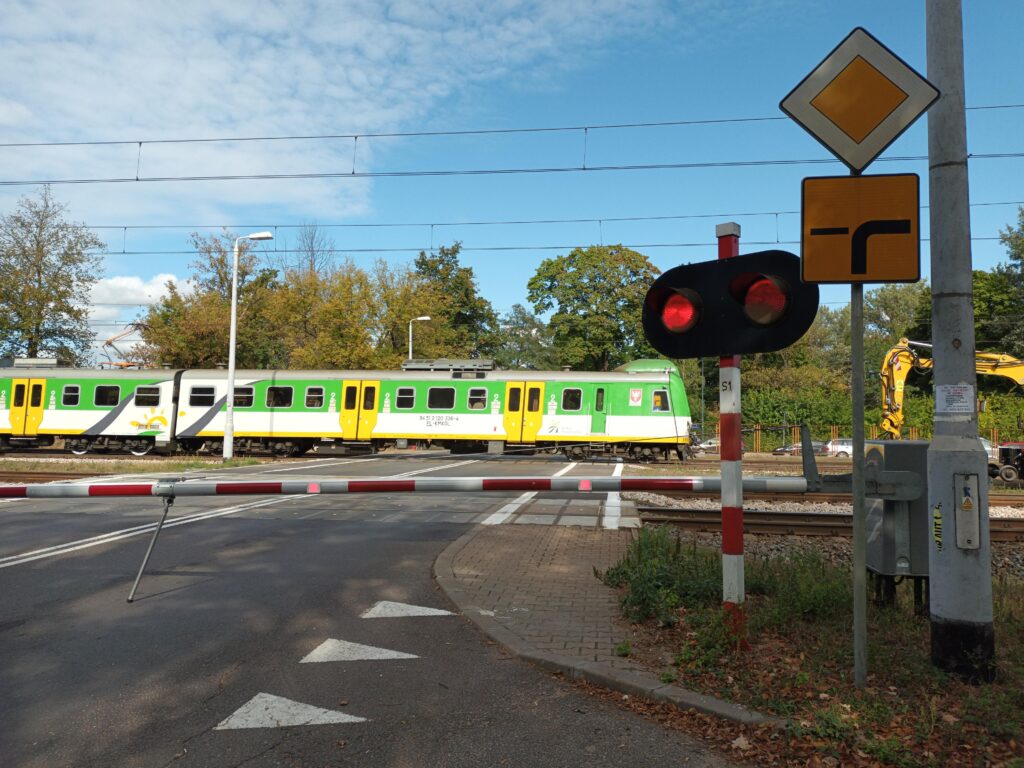 Pociąg w zielono-żółtej kolorystyce przejeżdża obok zamkniętej szlabanem drogi na przejeździe kolejowym.