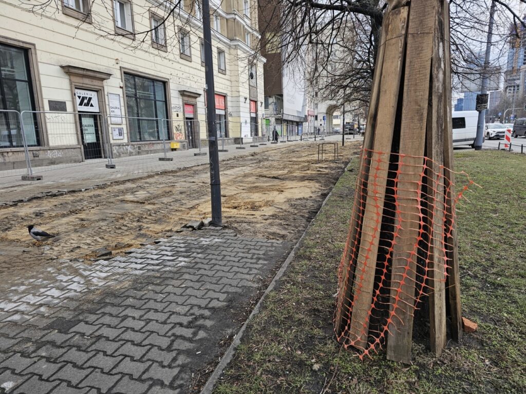 Pracownicy budowlani prowadzą prace remontowe na ulicy, odsłaniając podłoże i układając nowe kostki. W tle widać zamknięte sklepy.