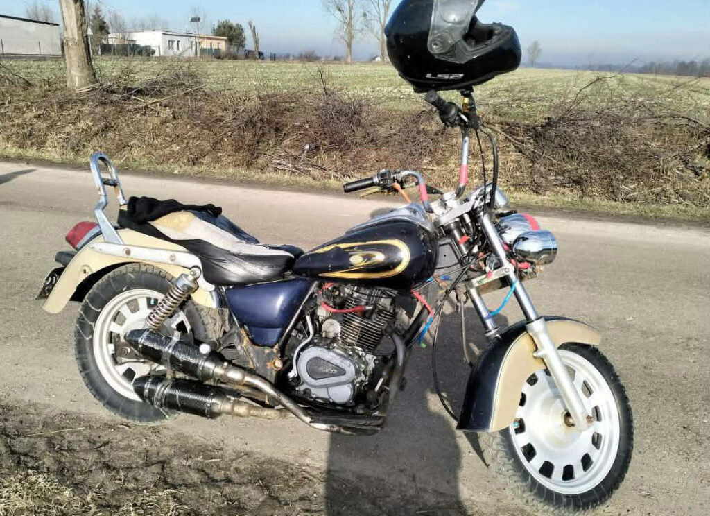 Motocykl z charakterystycznym, dużym przednim kołem stoi na poboczu drogi, otoczony przez pola i krzewy.