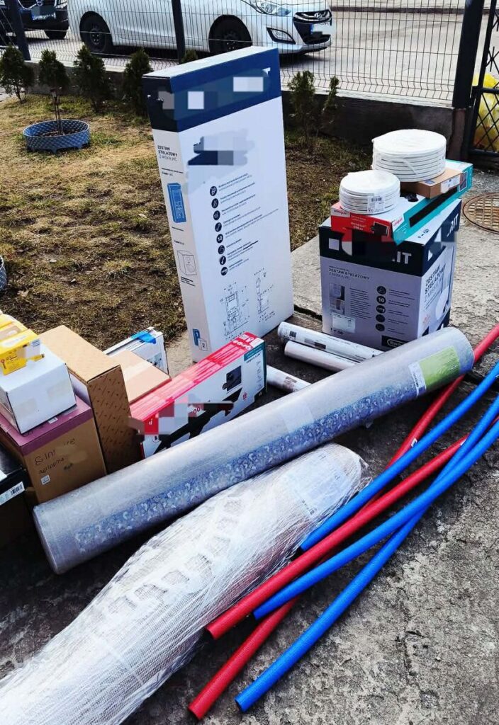 Na chodniku znajdują się różne materiały budowlane, w tym kartony, rury i rolety, obok trawnika.