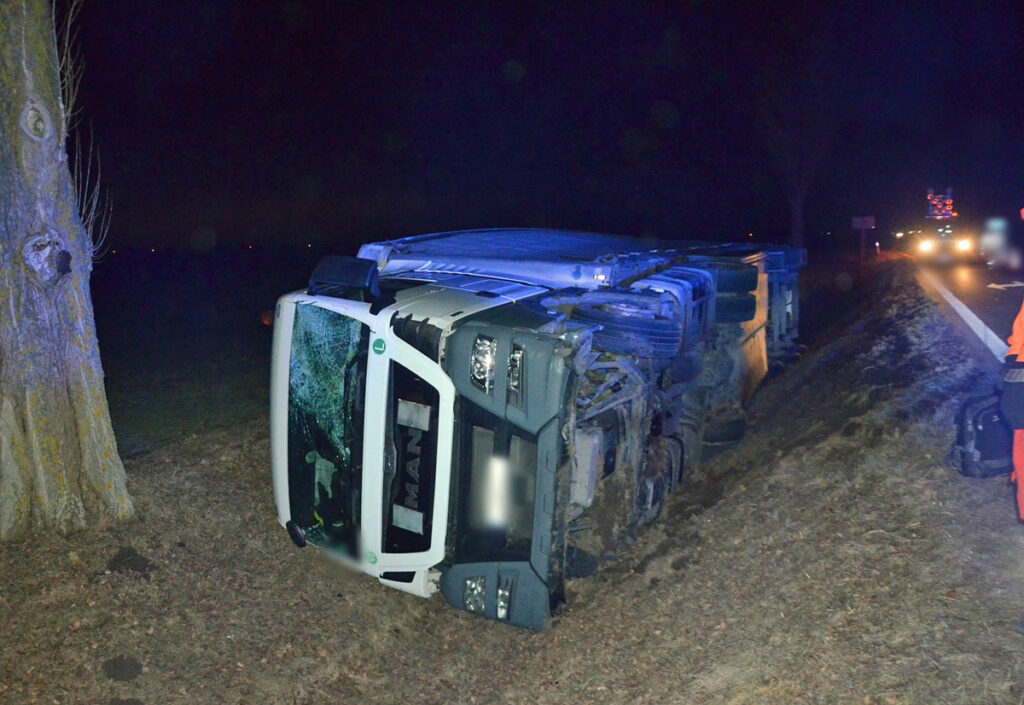 Przewrócony ciężarówka leży na boku przy drodze, obok interweniujący ratownik w pomarańczowym uniformie.