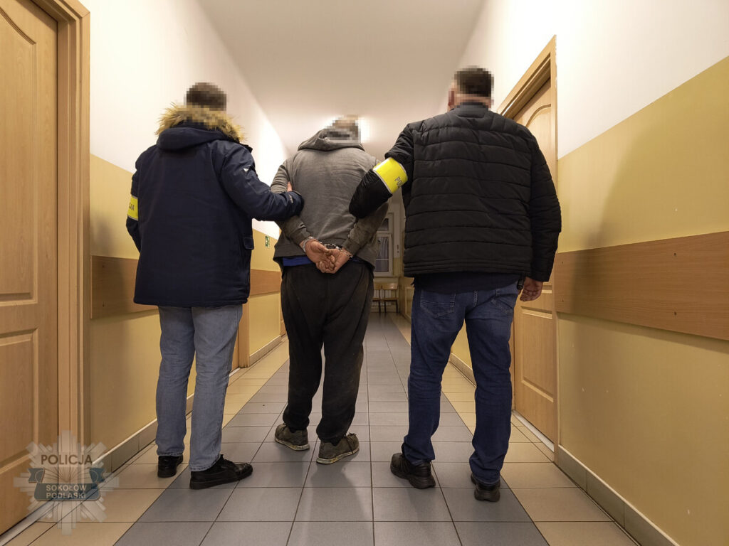 Trzej policjanci eskortują zatrzymanego mężczyznę korytarzem w budynku.