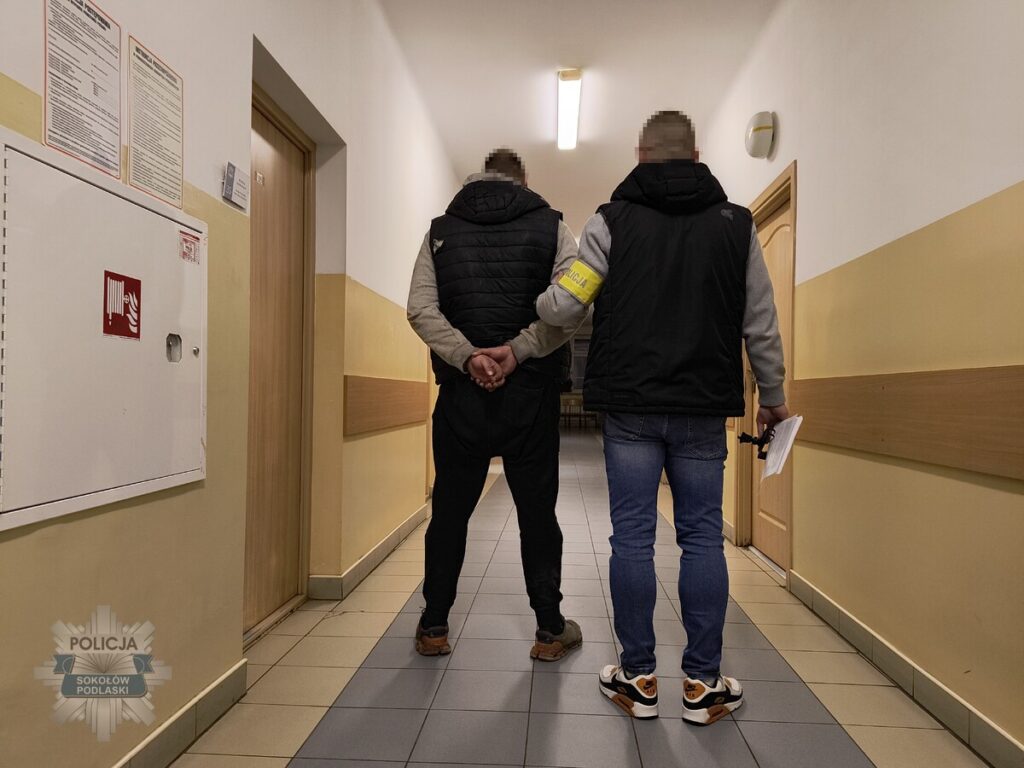 Mężczyzna z rękami skrępowanymi z tyłu prowadzony przez policjanta korytarzem w budynku publicznym.
