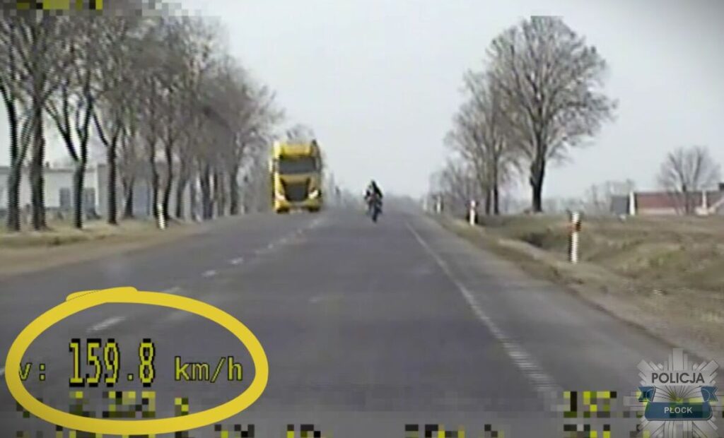Motocyklzysta porusza się z prędkością 159,8 km/h na drodze otoczonej drzewami. W tle widoczny żółty autobus.