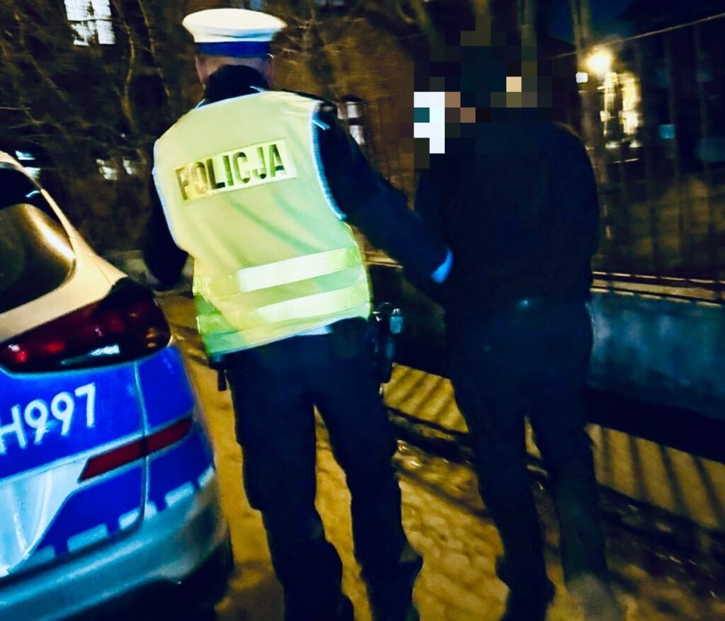 Policjant w kamizelce odblaskowej eskortuje zatrzymanego mężczyznę w nocy na ulicy.