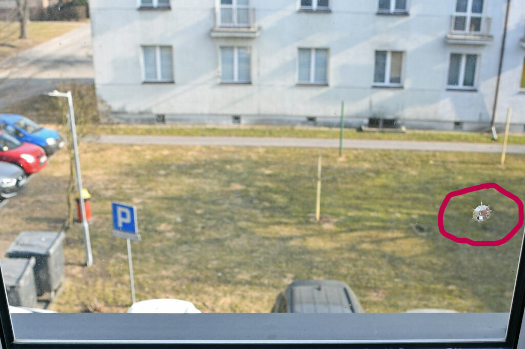 Na szybie widać niewielką dziurę, a w tle rozciąga się widok na parking z zaparkowanymi samochodami i trawnikiem.