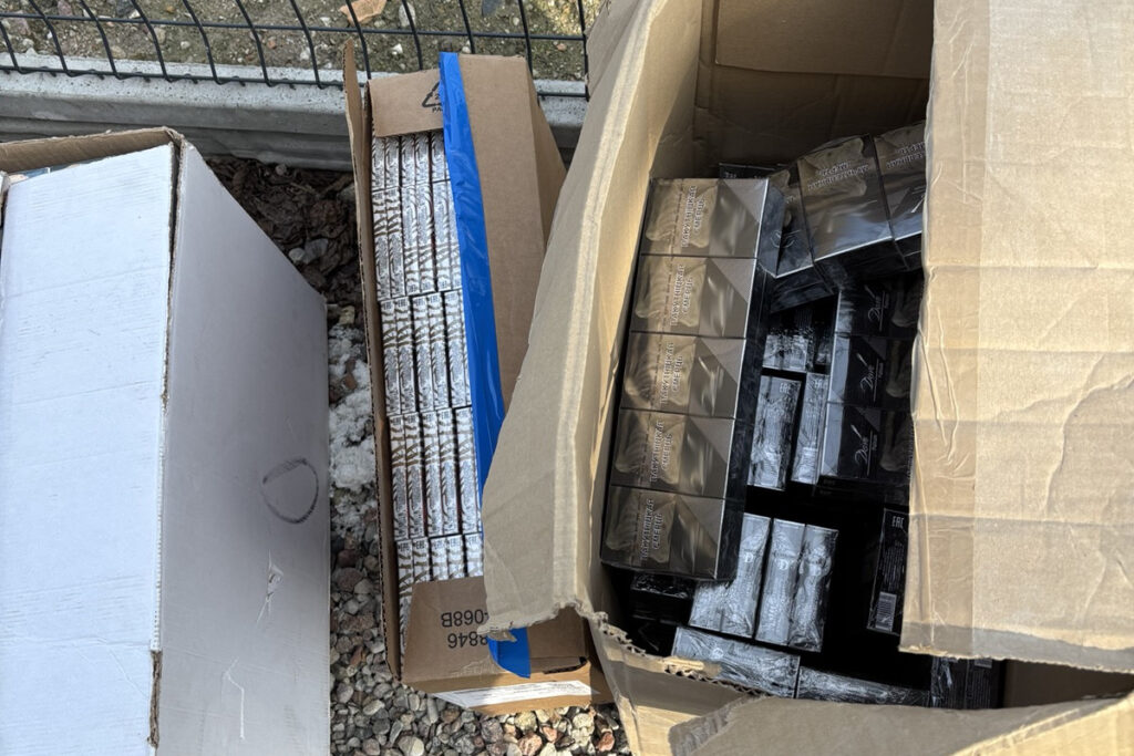 Dwa kartony z dużą ilością czarnych paczek, prawdopodobnie papierosów, leżą obok siebie na ziemi.