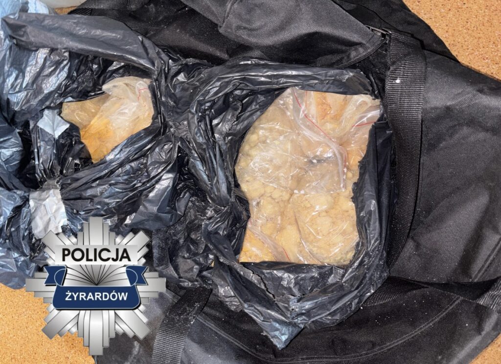 Czarna torba z przezroczystymi woreczkami zawierającymi nieznany materiał, leżąca na podłożu. W tle widoczny znak policji.