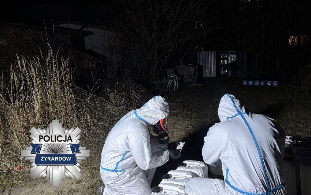 Dwaj funkcjonariusze w białych kombinezonach badają pojemniki w ciemnym otoczeniu, otoczeni wysoką trawą.