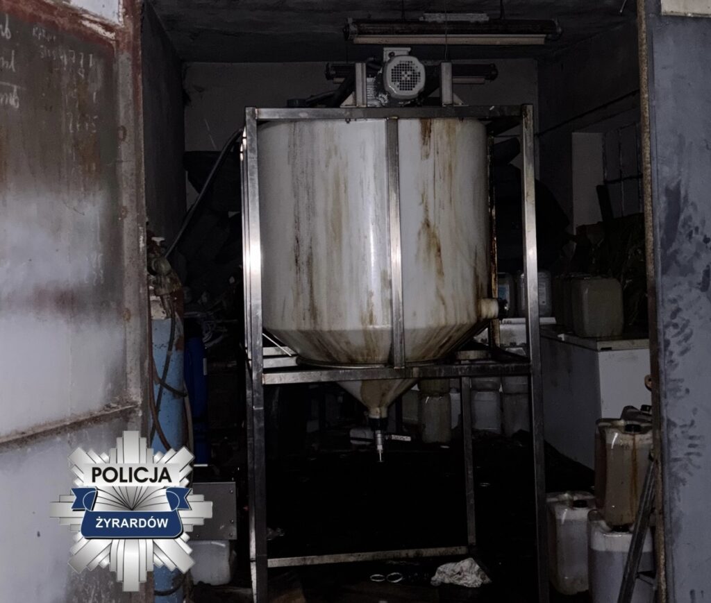 W pomieszczeniu widać metalowy zbiornik na stojaku, otoczony zanieczyszczonymi pojemnikami i ciemnym otoczeniem.