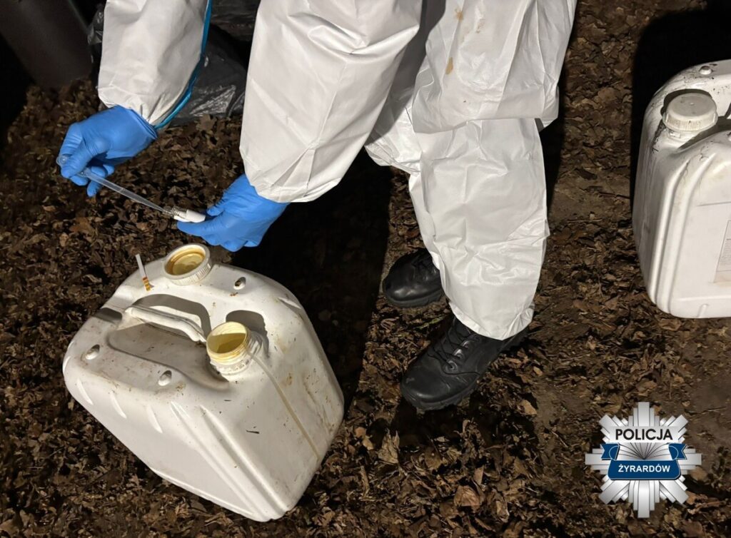 Osoba w białym kombinezonie ochronnym z niebieskimi rękawiczkami pobiera próbkę cieczy z plastikowego pojemnika na tle liści.