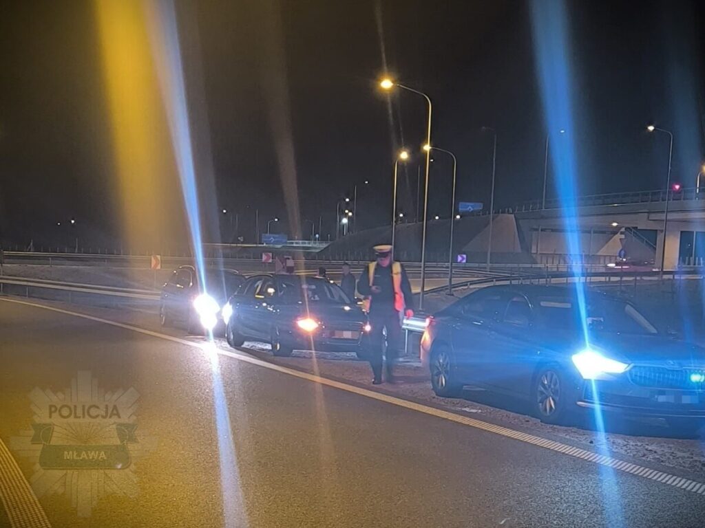 Policjant w odblaskowej kamizelce kontrolujący ruch na drodze w nocy, obok zatrzymane samochody.