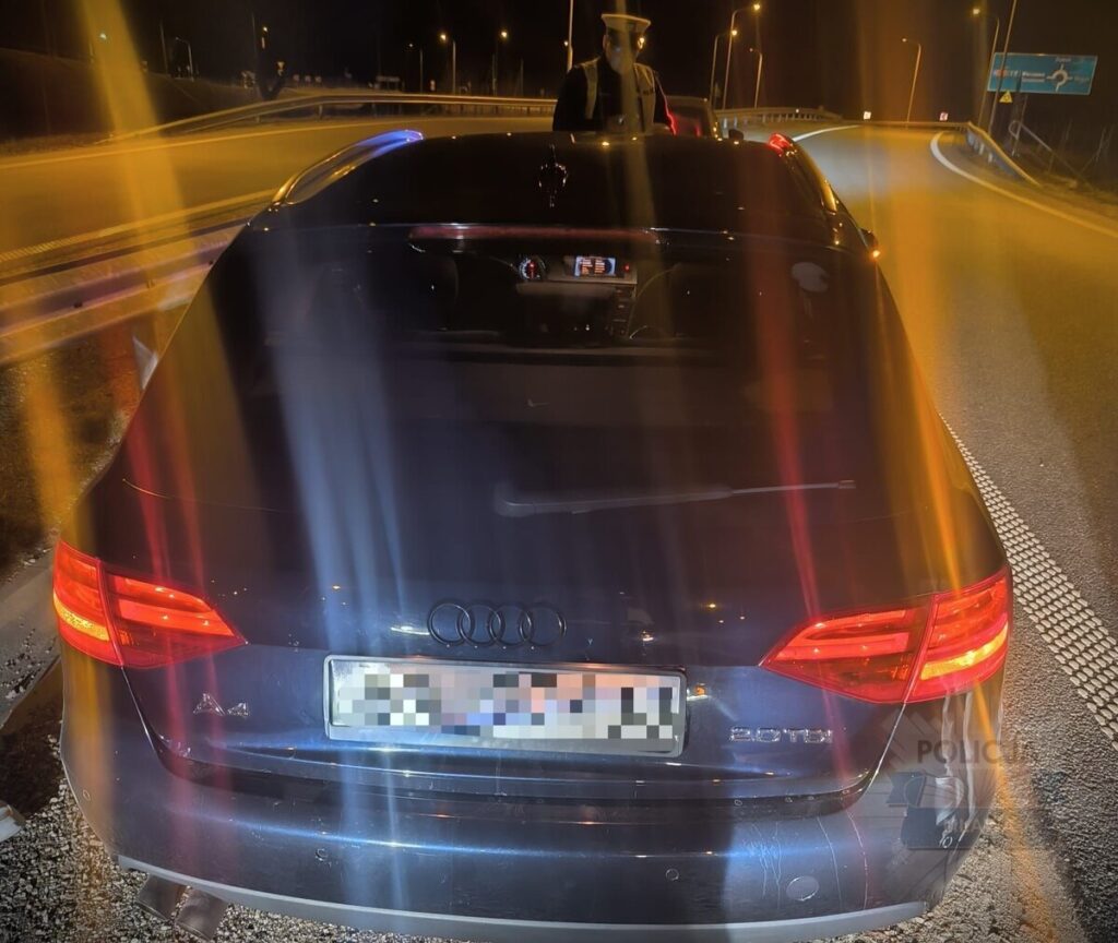 Policyjny funkcjonariusz stoi obok zatrzymanego samochodu osobowego marki Audi na drodze w nocy.