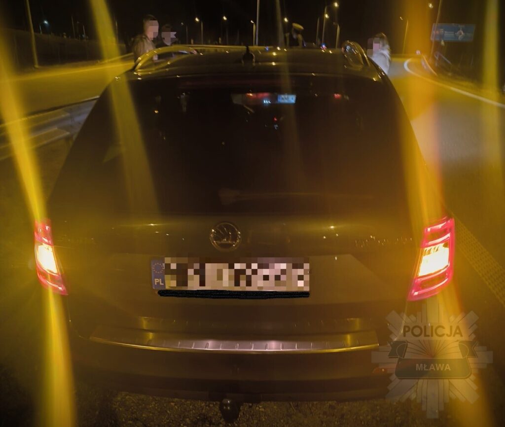Czarny samochód osobowy zatrzymany na drodze, w tle widoczni policjanci oraz oświetlenie uliczne w nocy.