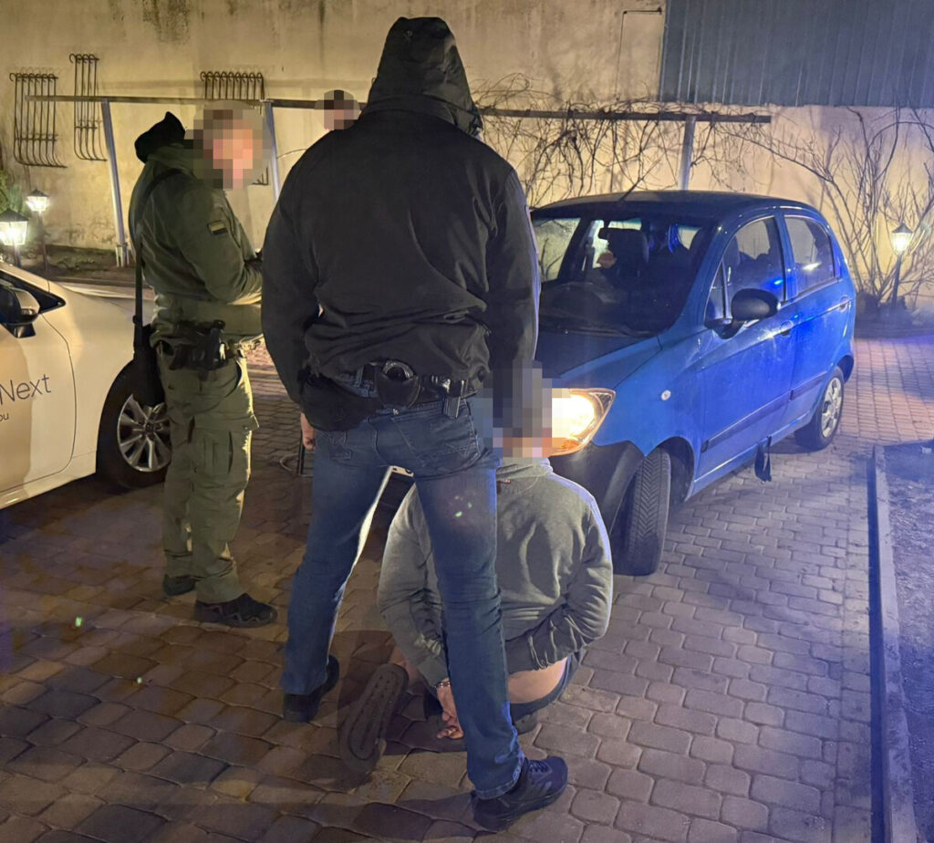 Funkcjonariusze policji zatrzymują mężczyznę na chodniku obok niebieskiego samochodu.