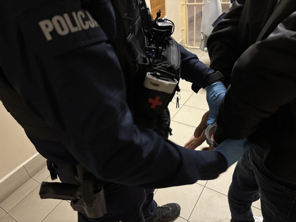 Policjant w mundurze zakłada kajdanki na ręce zatrzymanego mężczyzny w pomieszczeniu z kafelkową podłogą.