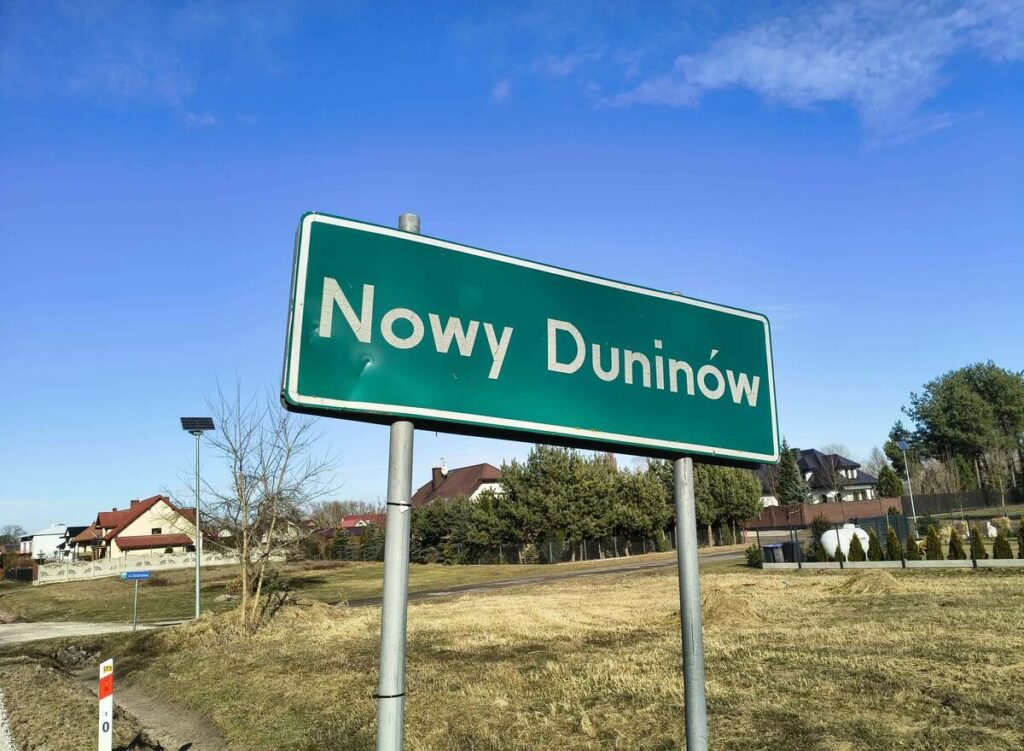 Zielony znak drogowy z napisem "Nowy Duninów" umieszczony przy asfaltowej drodze w otoczeniu domów i drzew.