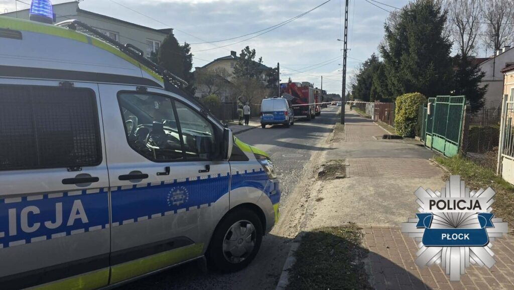 Radiowóz policyjny z włączonymi sygnałami świetlnymi stoi na ulicy, obok pojazdu strażackiego, w otoczeniu domów.