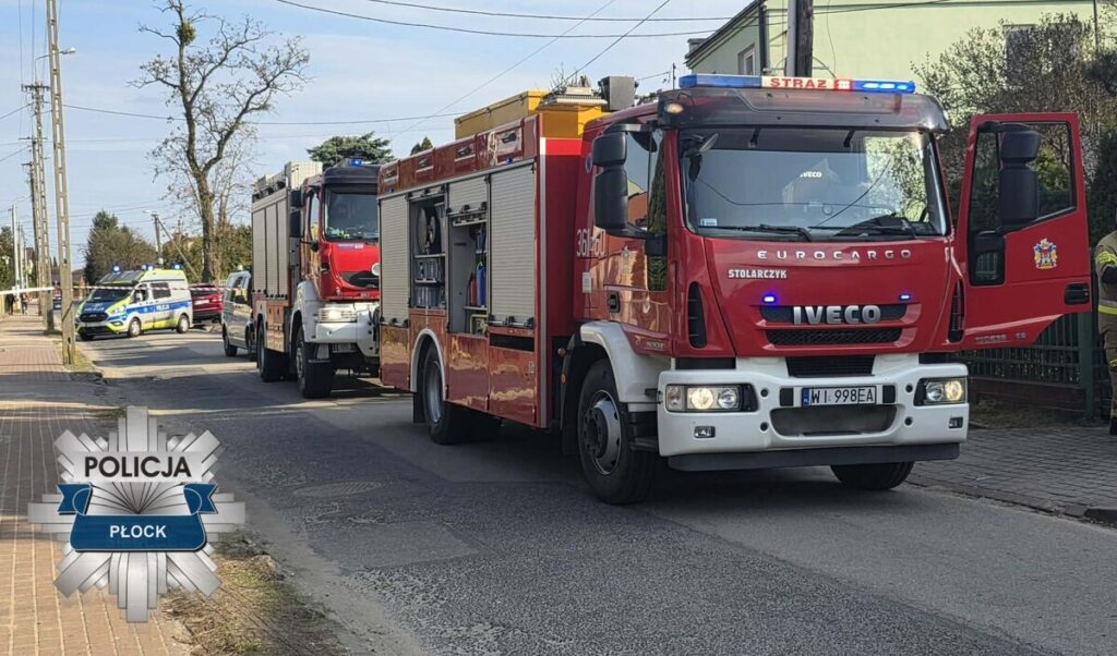 Czerwony wóz strażacki z otwartymi drzwiami stoi na ulicy, obok niego policyjny radiowóz oraz drugi samochód strażacki.