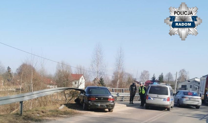 Czarny samochód osobowy zatrzymany przy barierce mostu, obok policjanci rozmawiający z kierowcą.