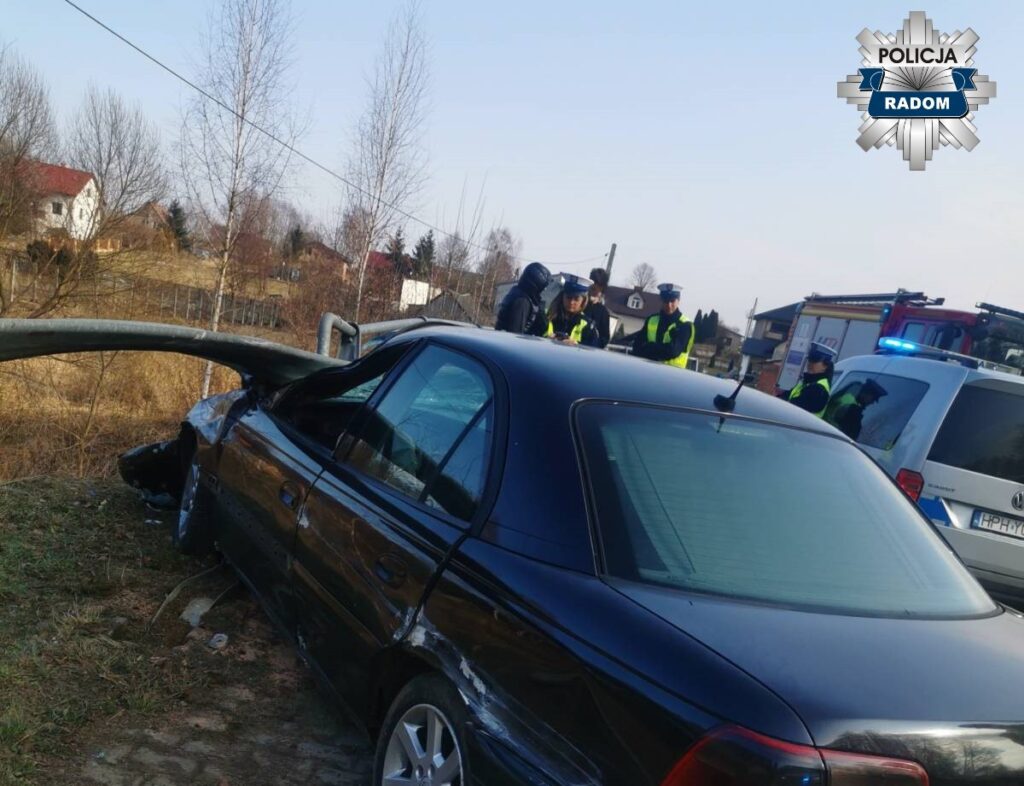 Czarny samochód osobowy z uszkodzeniami stoi obok przewróconego słupa, a policjanci prowadzą czynności na miejscu zdarzenia.
