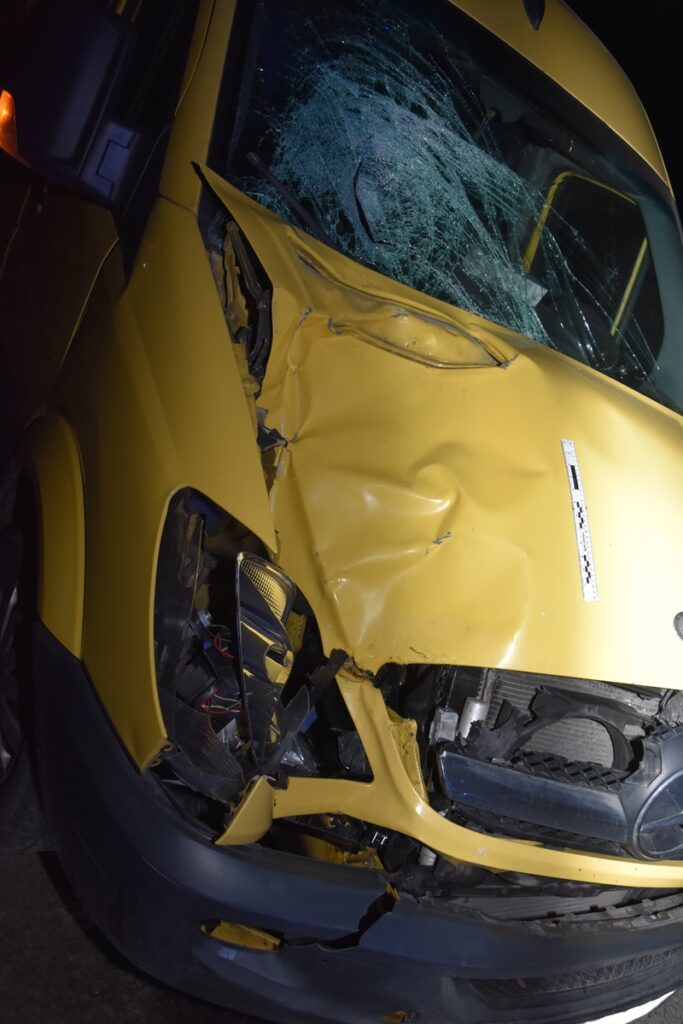 Żółty van z uszkodzoną przednią częścią oraz rozbitą szybą, stojący w ciemnym otoczeniu.