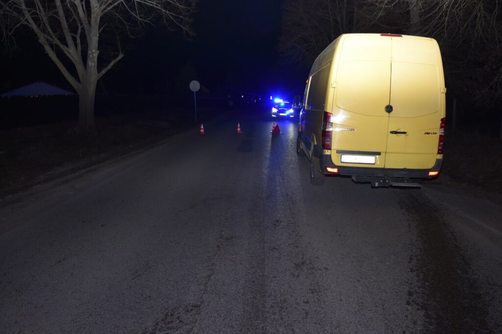 Żółty dostawczy samochód zaparkowany na drodze, obok włączone światła policyjnych pojazdów oraz pachołki.